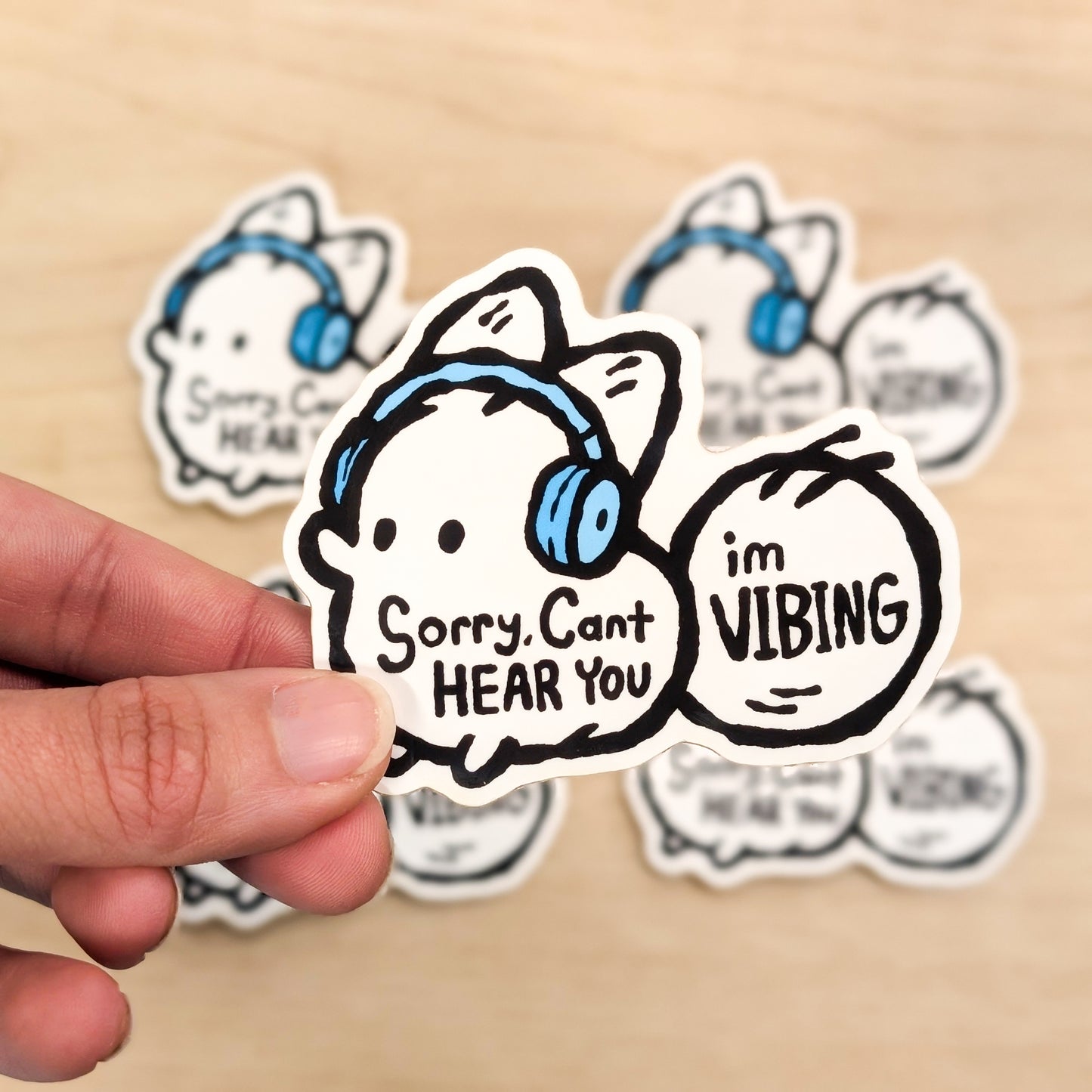 Sticker with cartoon character wearing headphones and text 'Sorry, Can't HEAR YOU I'm VIBING' held by a hand on a beige background.