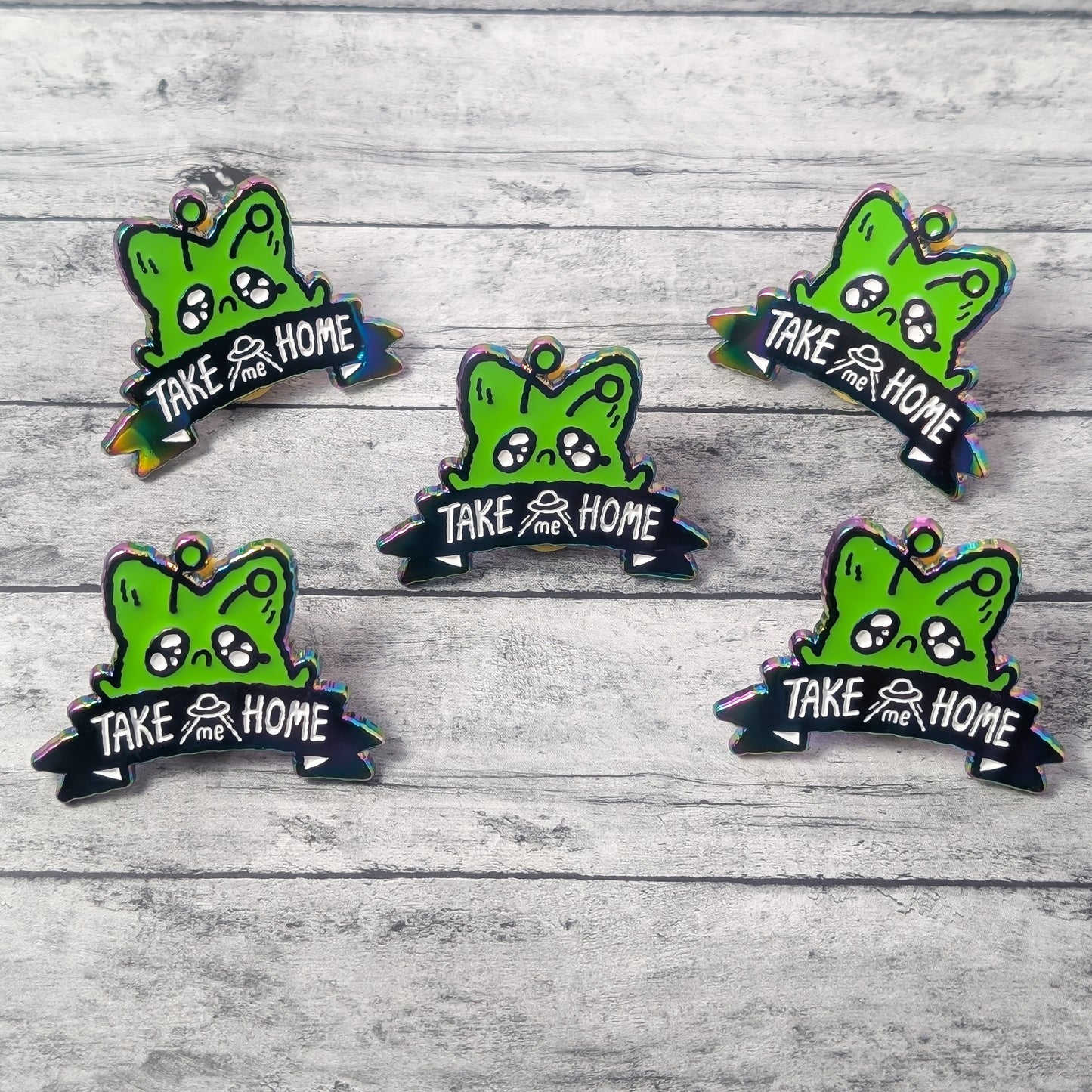 Five green cartoon alien fox pins with 'Take me Home' text on a wooden surface