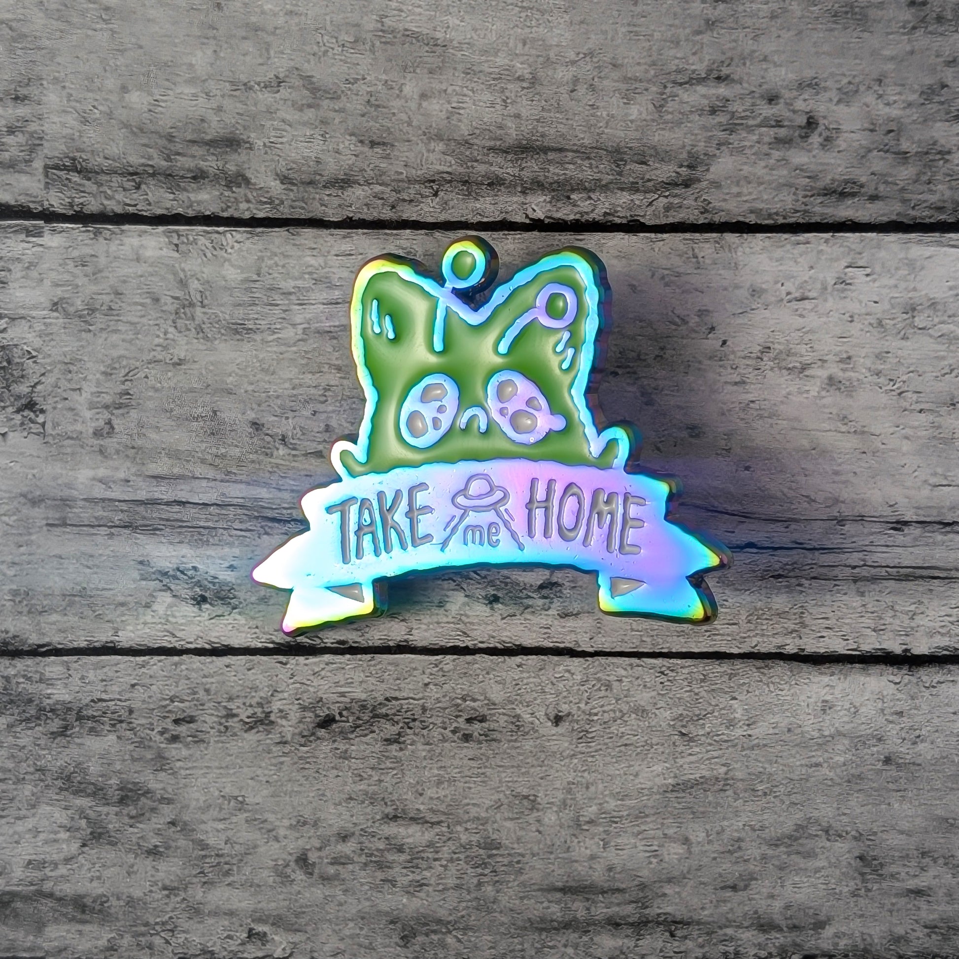 Enamel pin with a green alien fox and 'Take me Home' text on a wooden background