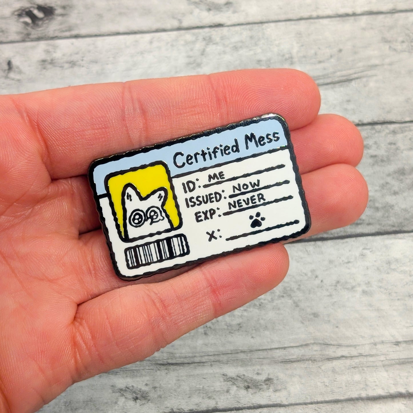 Hand holding an enamel pin with a humorous 'Certified Mess' design on a wooden surface