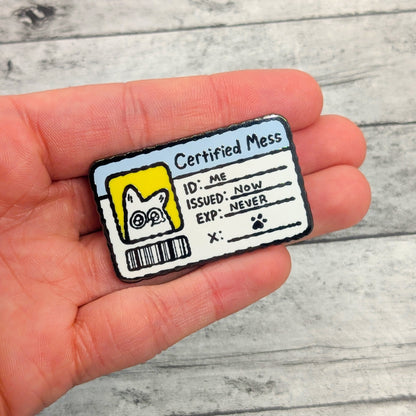 Hand holding an enamel pin with a humorous 'Certified Mess' design on a wooden surface