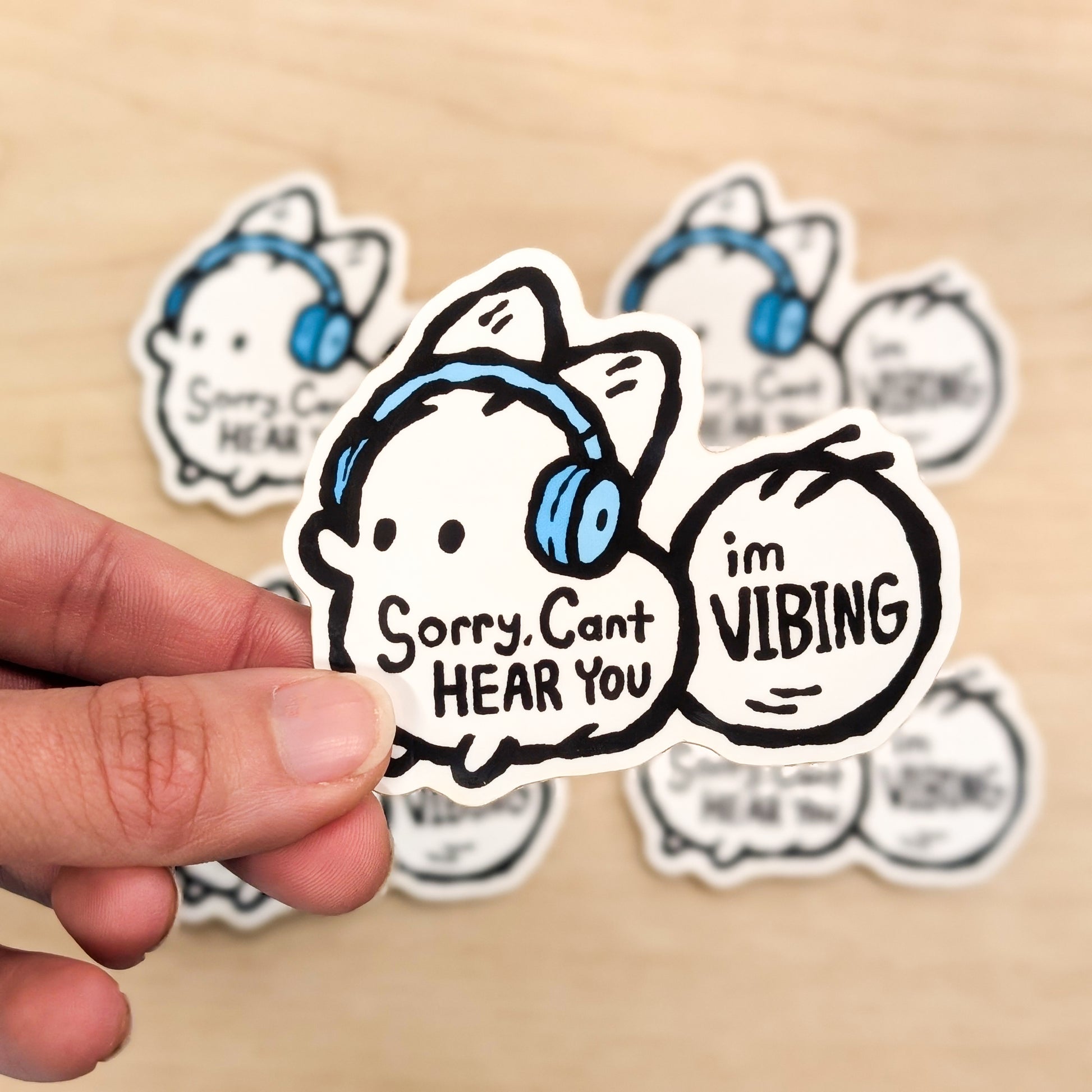Sticker with cartoon character wearing headphones and text 'Sorry, Can't HEAR YOU I'm VIBING' held by a hand on a beige background.