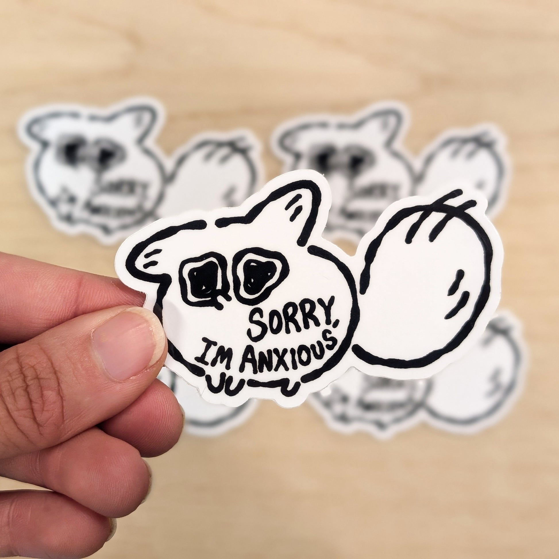 Sticker with a white fox and 'Sorry, I'm Anxious' text held by a hand on a beige background.