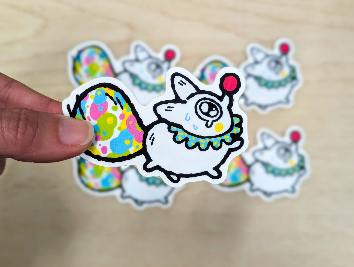 Hand holding a colorful sticker of a cartoon white fox with a clown outfit on a wooden surface.