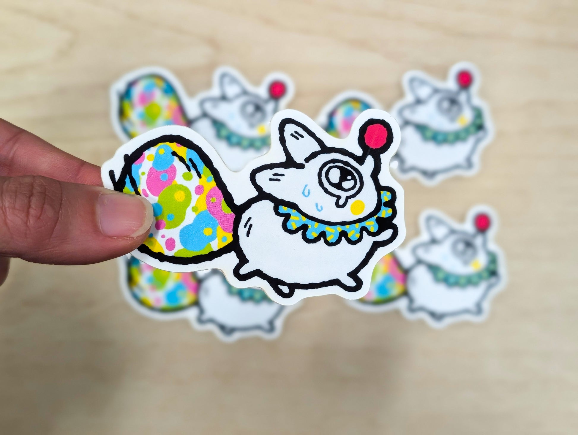 Hand holding a colorful sticker of a cartoon white fox with a clown outfit on a wooden surface.