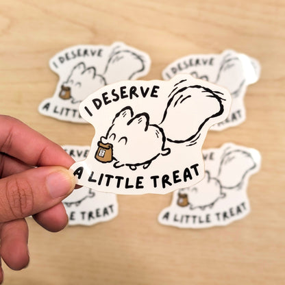Hand holding a sticker with a cartoon white fox and text 'I DESERVE A LITTLE TREAT' on a wooden surface.