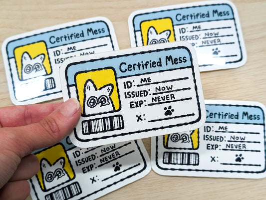 Hand holding a 'Certified Mess' ID vinyl sticker with multiple stickers on a wooden surface