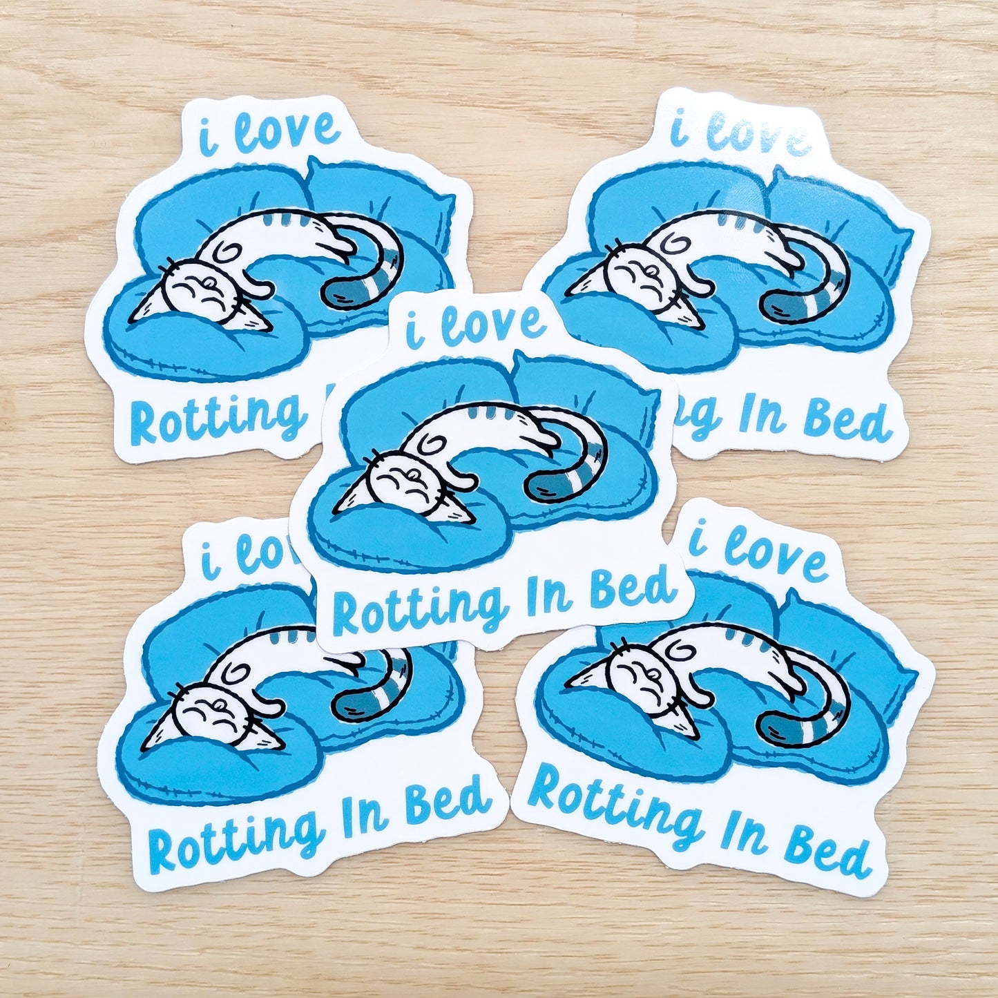 Five stickers with a cat design and text 'i love Rotting In Bed' on a wooden surface.