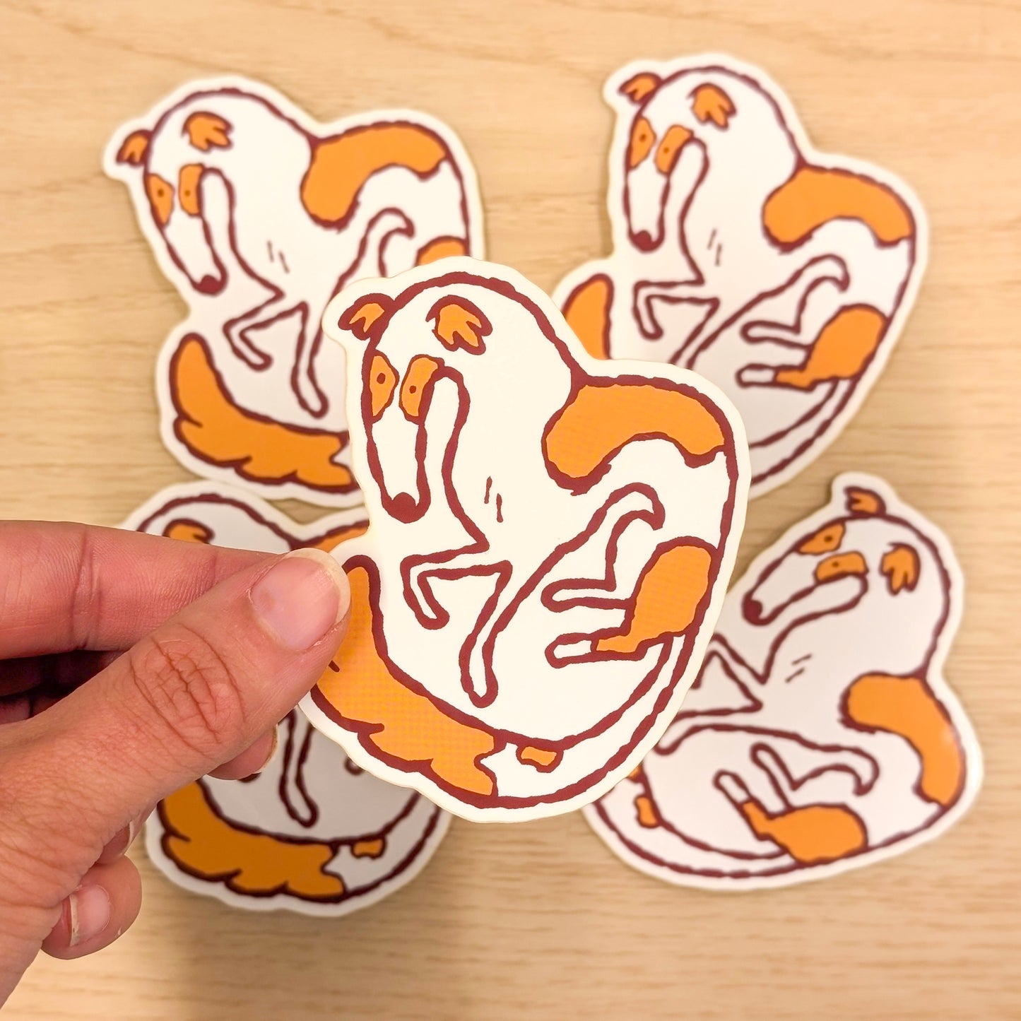 Hand holding a sticker of a Borzoi with orange and white design on a wooden surface