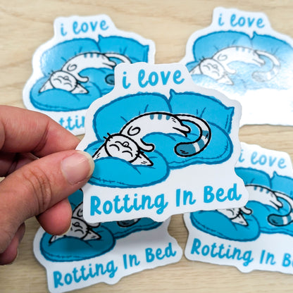 Sticker with a cat on a blue blanket and text 'i love rotting in bed' held by a hand.