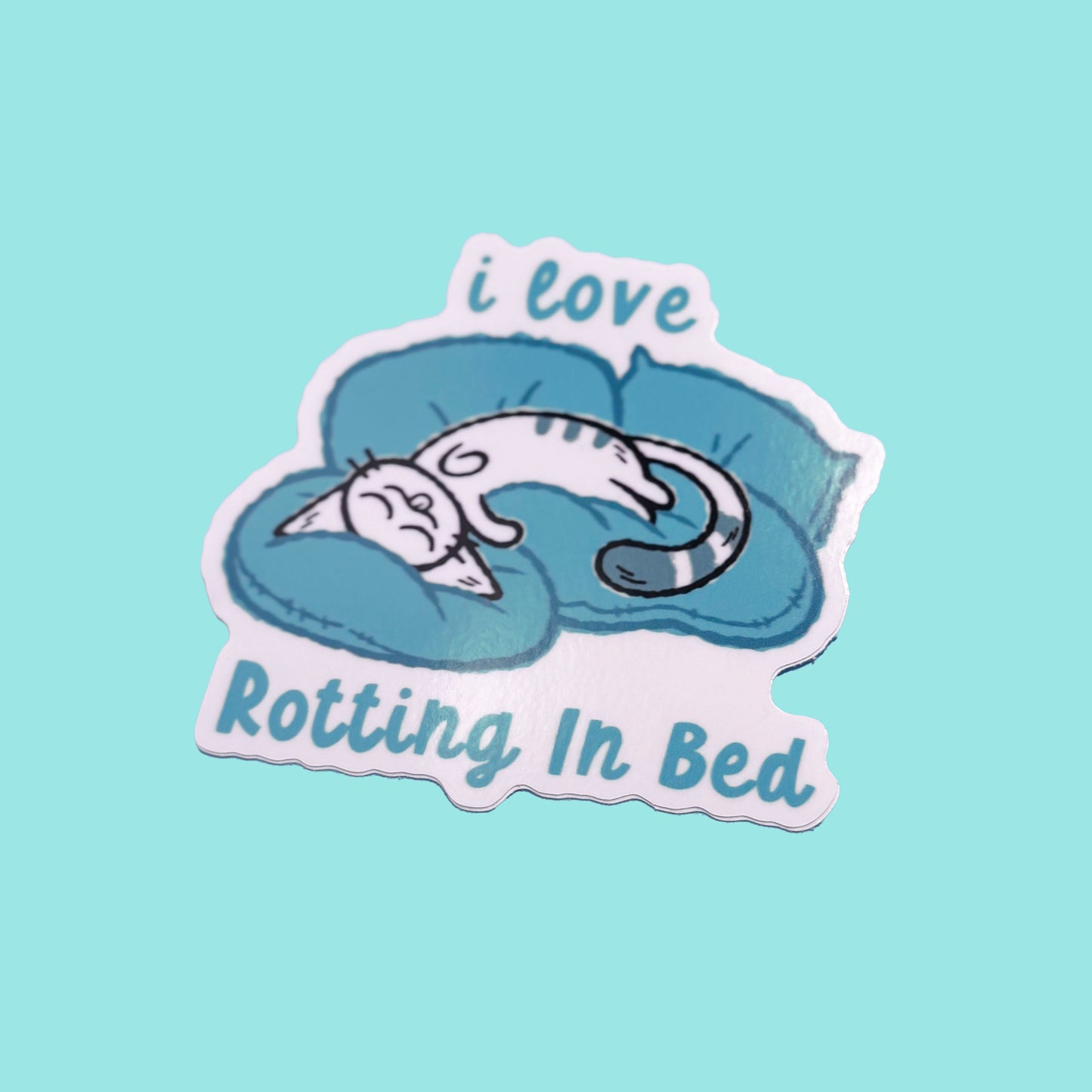 Sticker with a cat sleeping on a pillow and text 'i love Rotting In Bed' on a light blue background