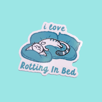 Sticker with a cat sleeping on a pillow and text 'i love Rotting In Bed' on a light blue background