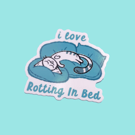 Sticker with a cat sleeping on a pillow and text 'i love Rotting In Bed' on a light blue background