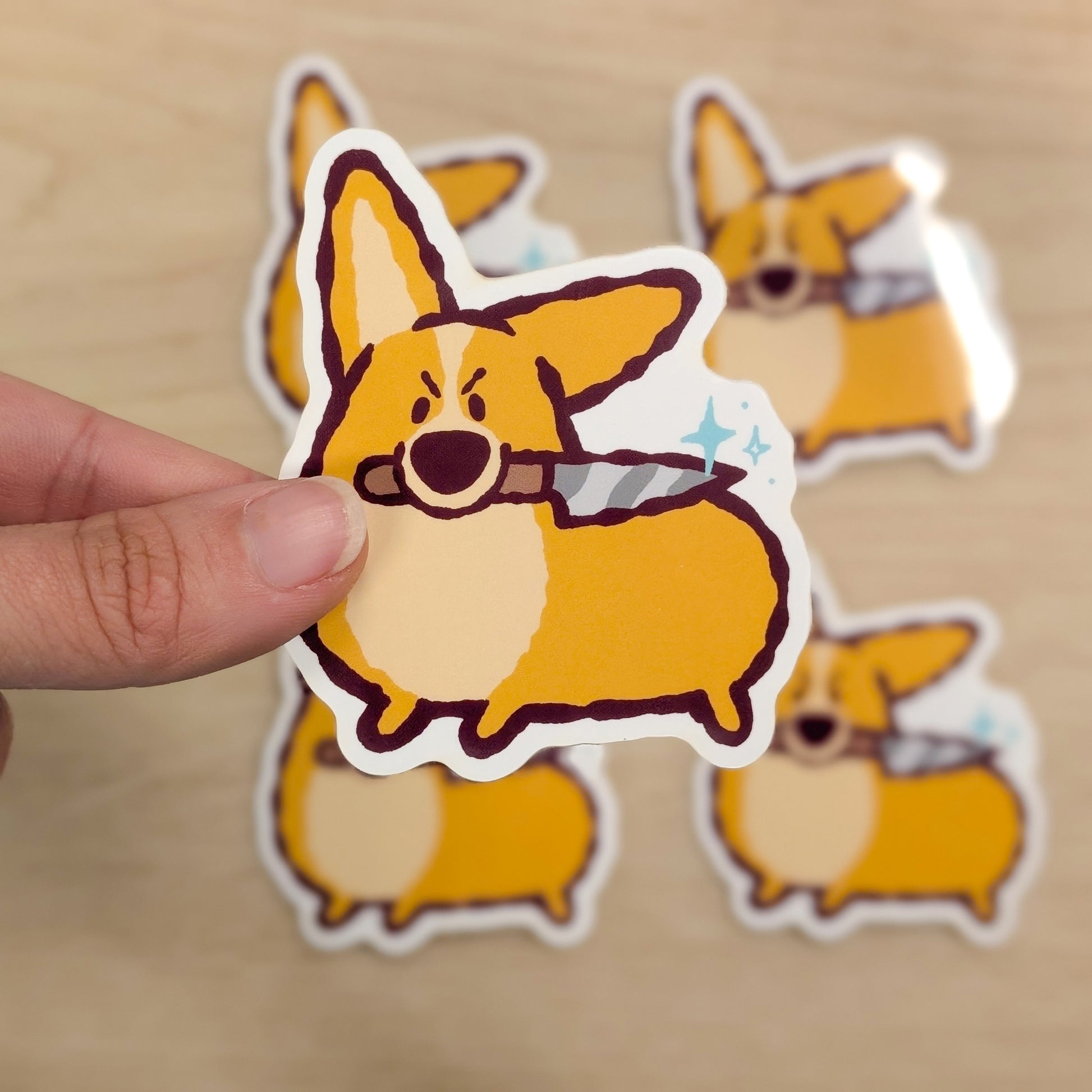 Hand holding a sticker of a corgi with a knife, with more stickers on a beige background
