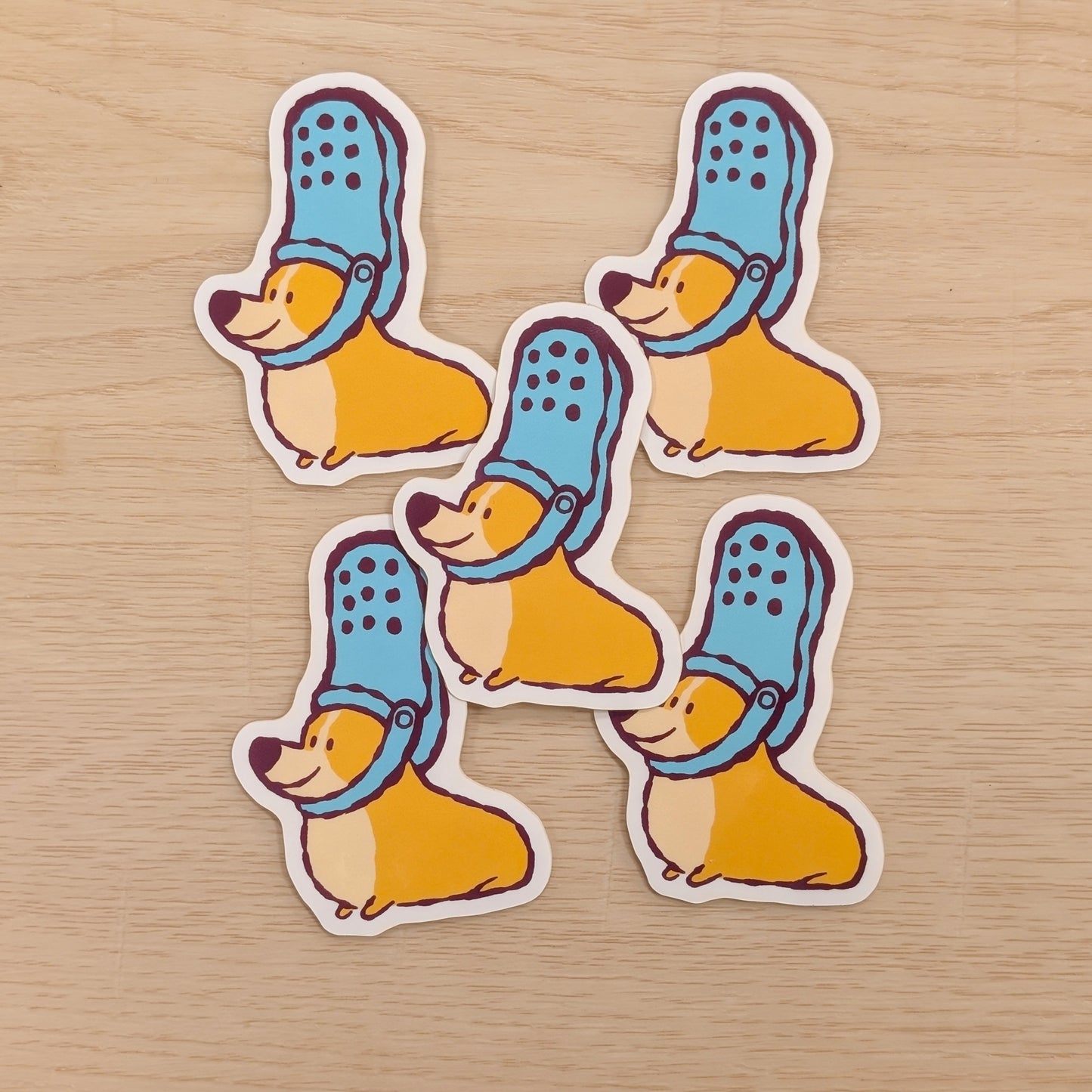 Five stickers of corgis wearing blue shoes on their heads, on a wooden surface
