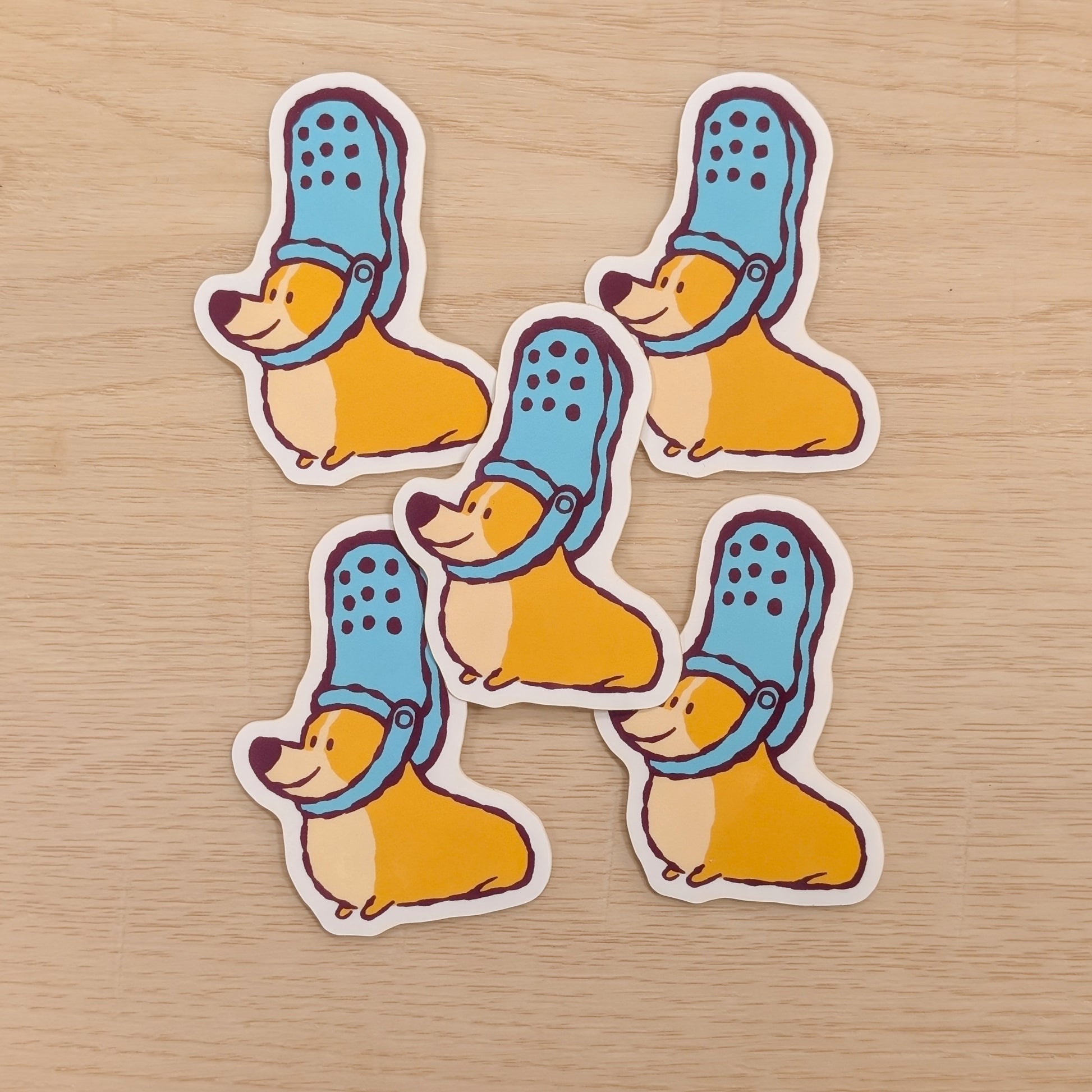 Five stickers of corgis wearing blue shoes on their heads, on a wooden surface