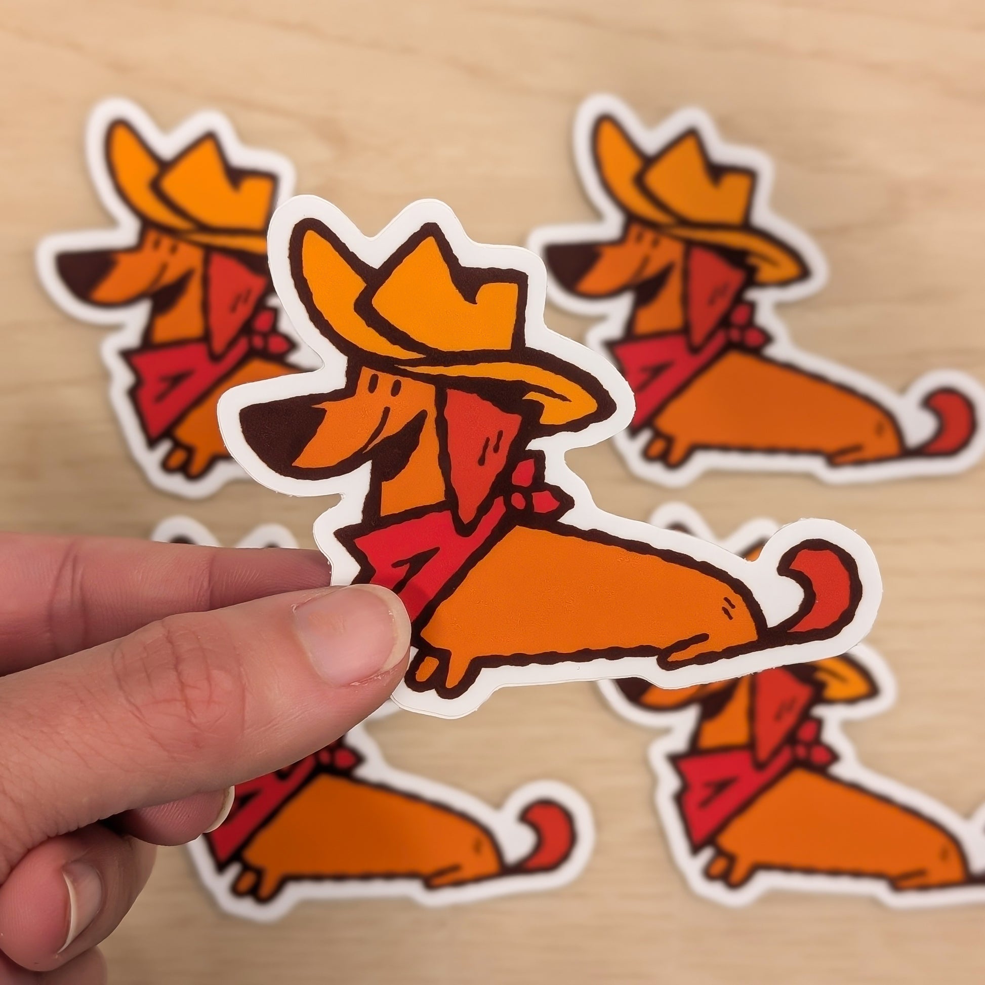 Hand holding a cowboy-themed sticker of a Dachshund dog on a wooden surface with more stickers in the background.