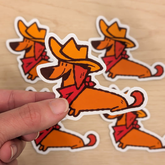 Hand holding a cowboy-themed sticker of a Dachshund dog on a wooden surface with more stickers in the background.