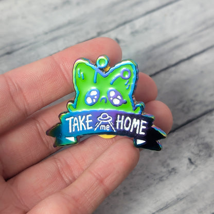 Colorful 'Take Me Home' pin featuring a green alien fox, held in a hand on a wooden surface