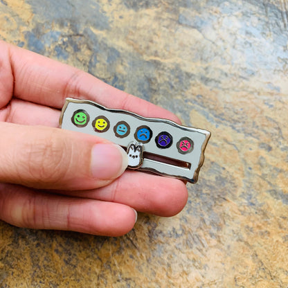 A hand holding a mood tracker enamel pin on a stone background