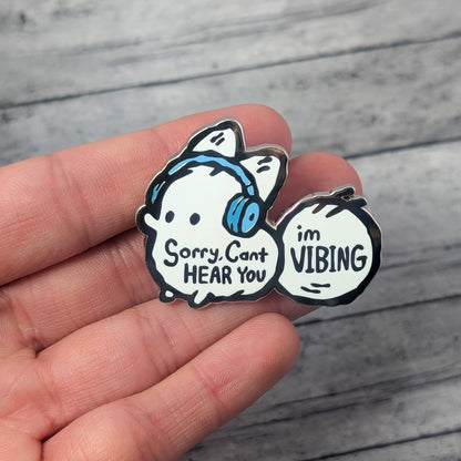 Hand holding an enamel pin of a white cartoon fox wearing headphones that says 'Sorry, can't hear you, I'm vibing.'