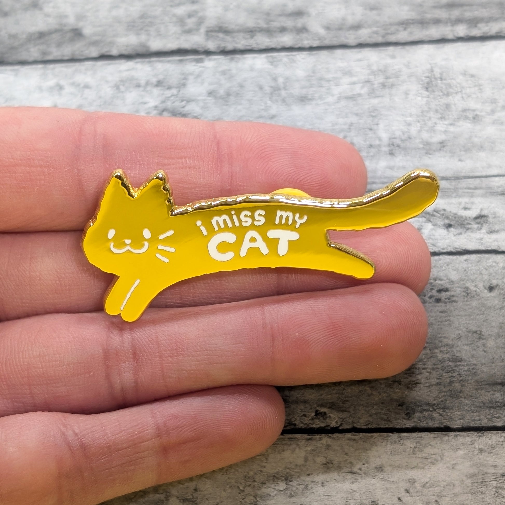 Yellow cat-shaped pin with 'I miss my CAT' text held between fingers on a wooden surface