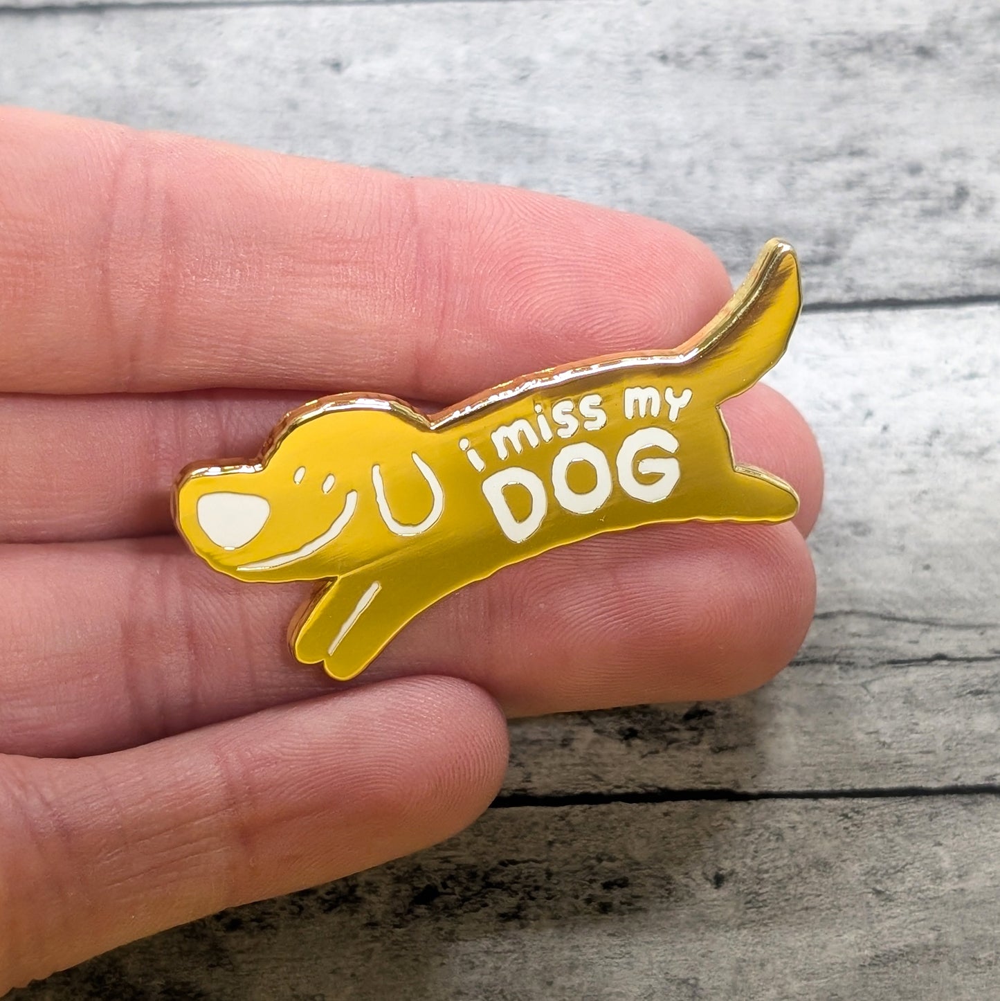 Yellow dog-shaped pin with 'I miss my DOG' text held in a hand on a wooden surface