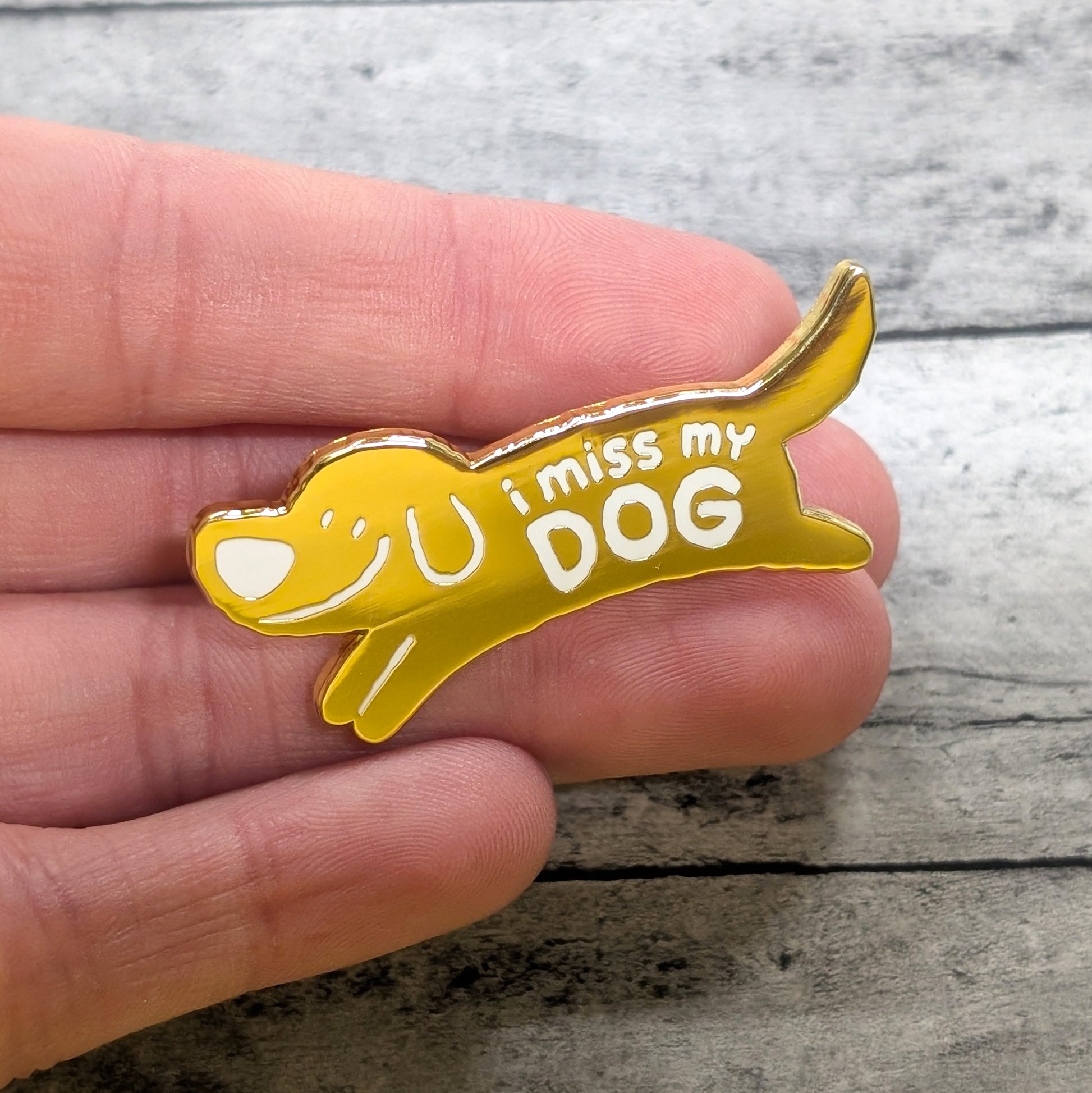 Yellow dog-shaped pin with 'I miss my DOG' text held in a hand on a wooden surface
