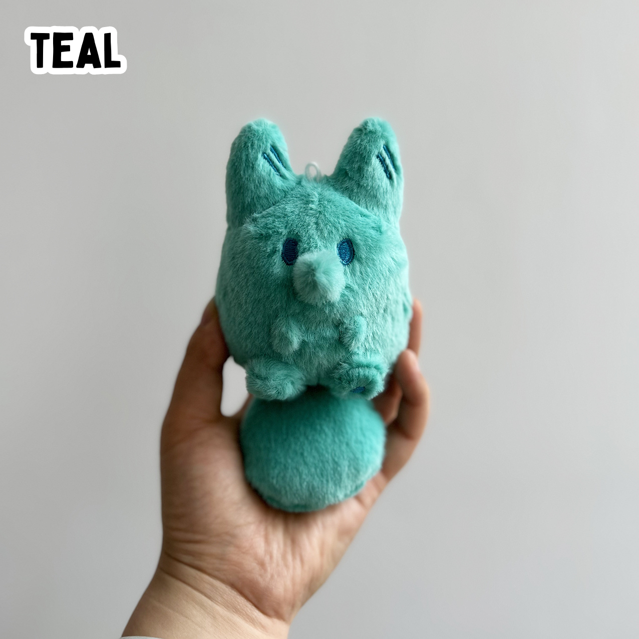 A hand holding a mini teal fox plush toy against a white background