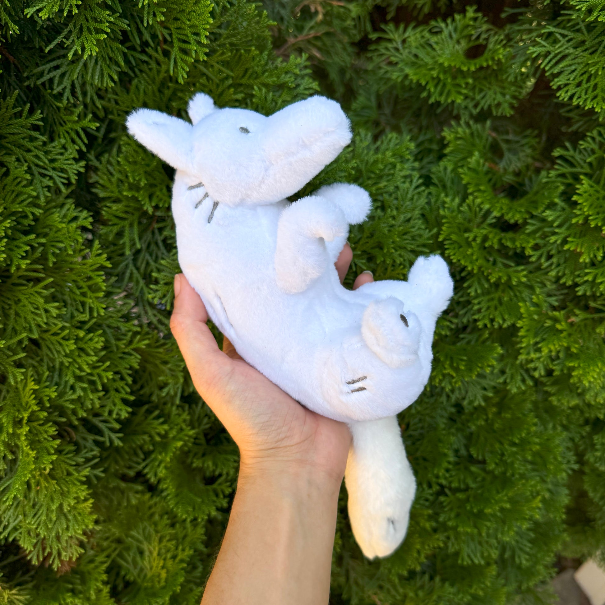 A hand holding a plush toy of a white wolf, with a bush in the background