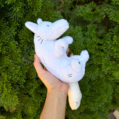 A hand holding a plush toy of a white wolf, with a bush in the background