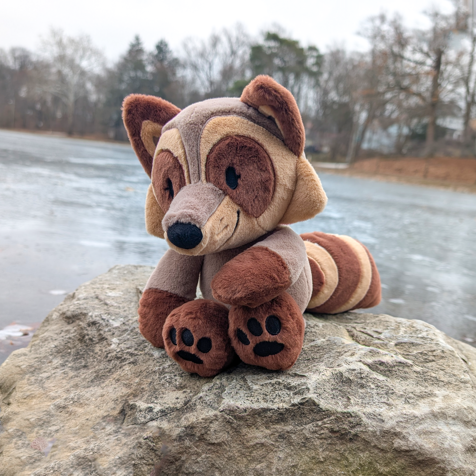 Plush toy of a brown raccoon sitting on a rock overlooking some water.