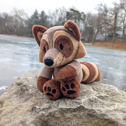 Plush toy of a brown raccoon sitting on a rock overlooking some water.