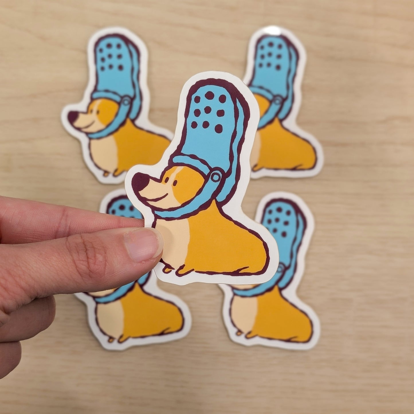 Sticker of a corgi wearing blue shoes on its head, held by a hand on a wooden surface, with more stickers in the background