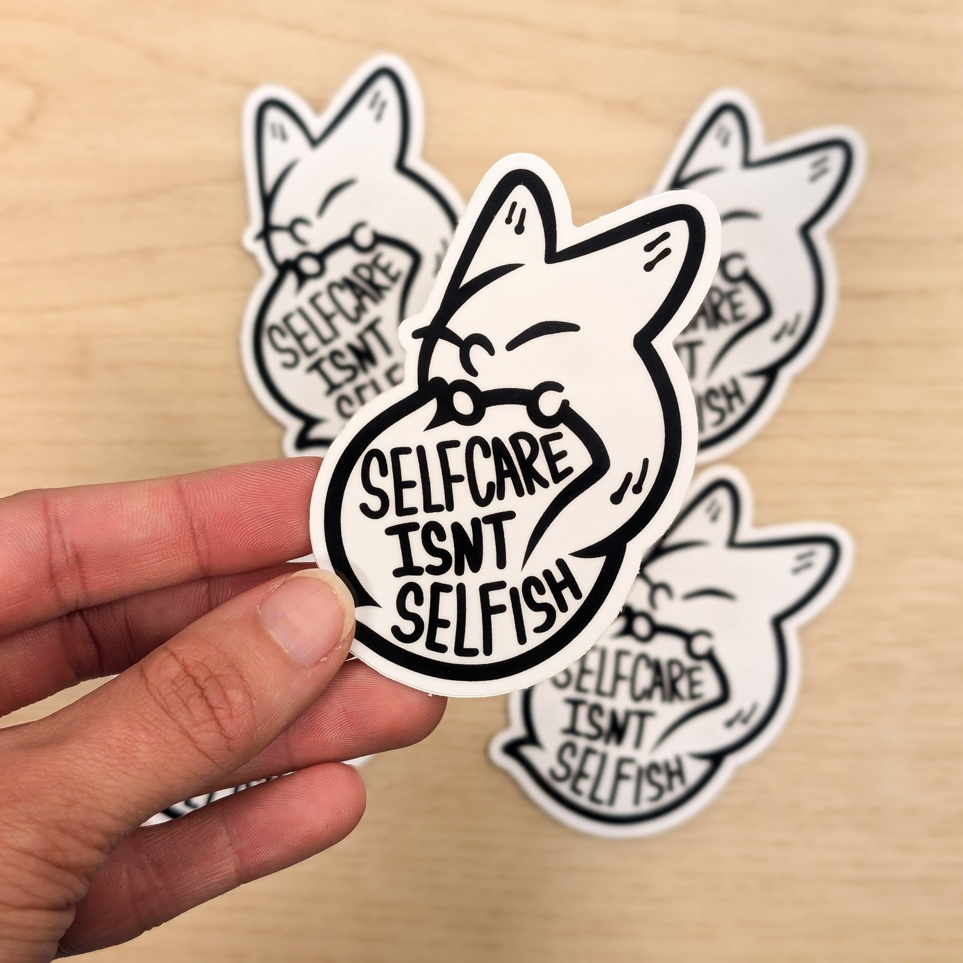 Hand holding a 'Self care Isn't Selfish' sticker with more stickers in the background on a wooden surface.
