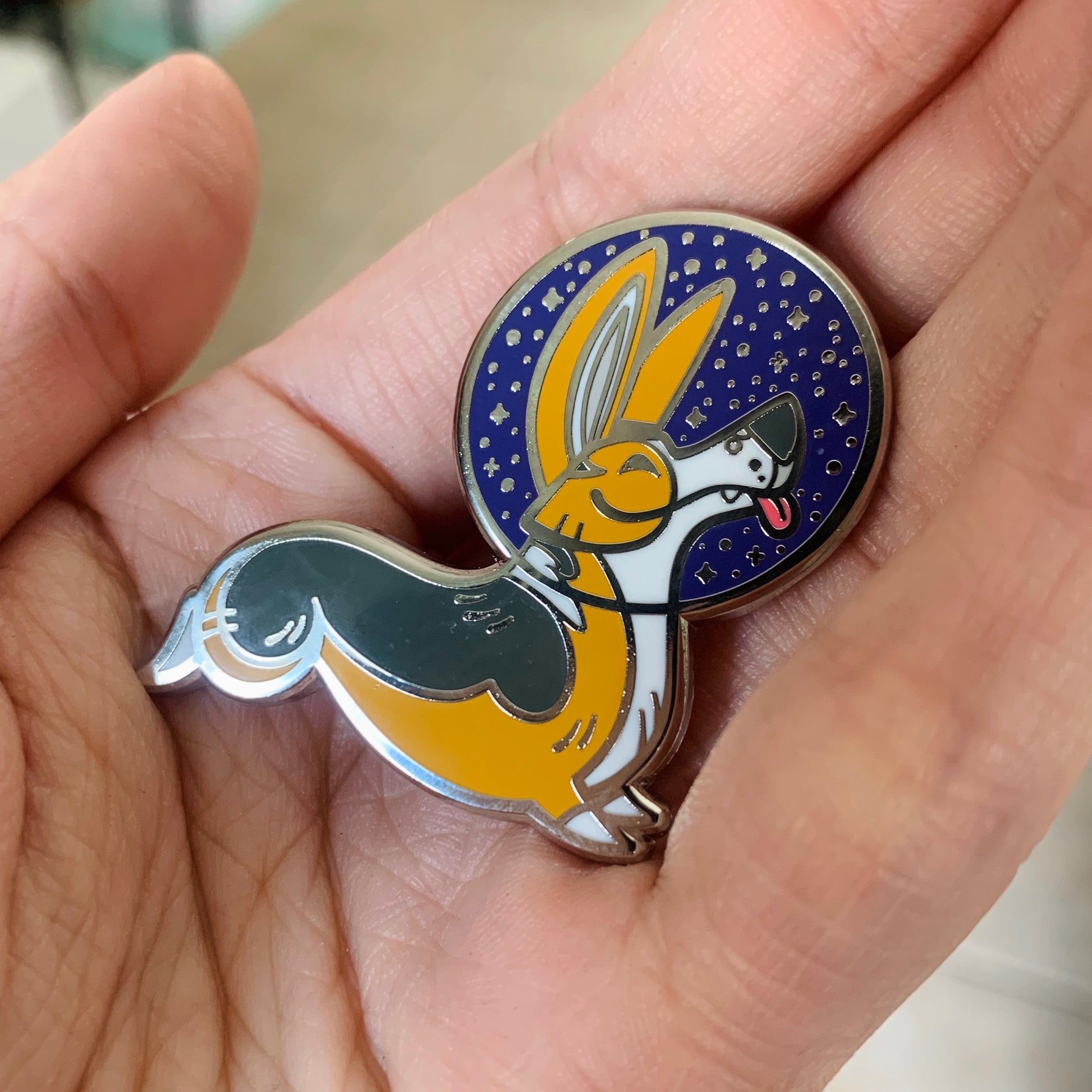 Hand holding an enamel pin with a stylized corgi wearing a space helmet.